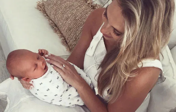 A woman holding a swaddled newborn baby on a couch, looking down at the child lovingly.