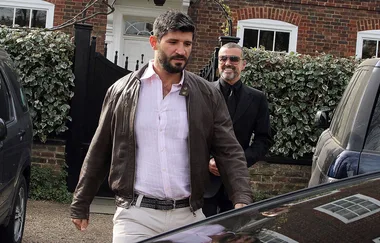 Two men walking near parked cars in front of a house with a brick exterior and white trim.