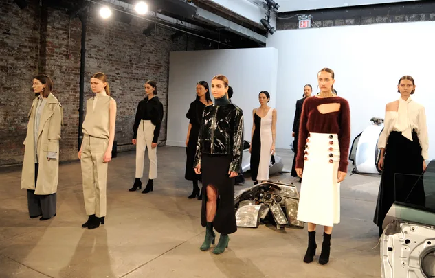Models in diverse outfits stand in an industrial-style space with exposed brick walls during a fashion presentation.
