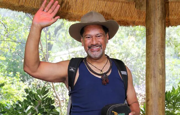A person in a hat and tank top smiles and waves in a natural setting.