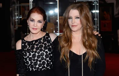 Two women pose together at an event, one wearing a black polka dot dress, the other in a black outfit with pearls.