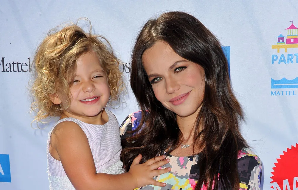 Two people smiling, one is a young child with curly hair, the other has long dark hair, against a blue event backdrop.