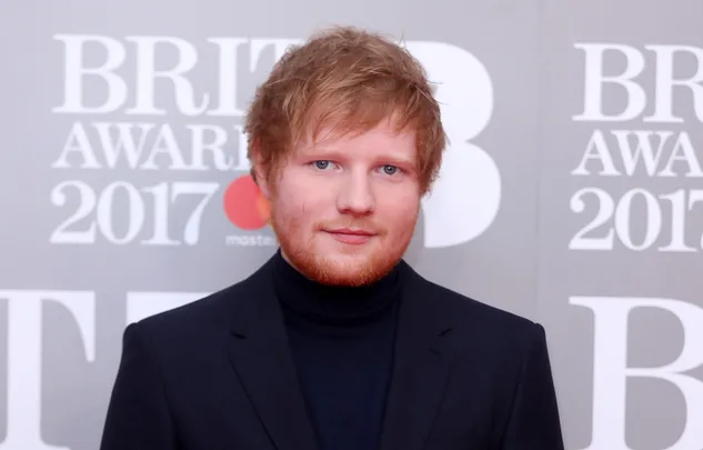 Red-haired person in a black suit at the BRIT Awards 2017, standing in front of a sign with red lettering.