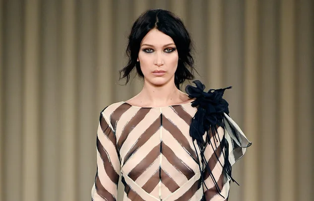 A model walks the runway wearing a sheer striped dress with a black floral accent at the shoulder, against a beige background.
