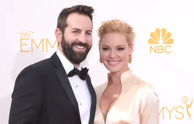 Couple smiling on the red carpet at the 66th Emmy Awards, with NBC and Emmy logos in the background.
