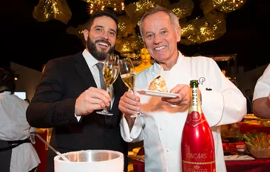 Two chefs toasting with champagne, holding dessert near an Oscar statue, with festive decorations in the background.