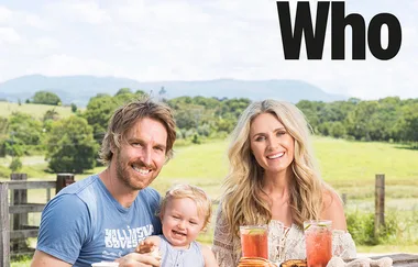 A smiling couple with a child sit outdoors with drinks and food on a picnic table; countryside in the background.