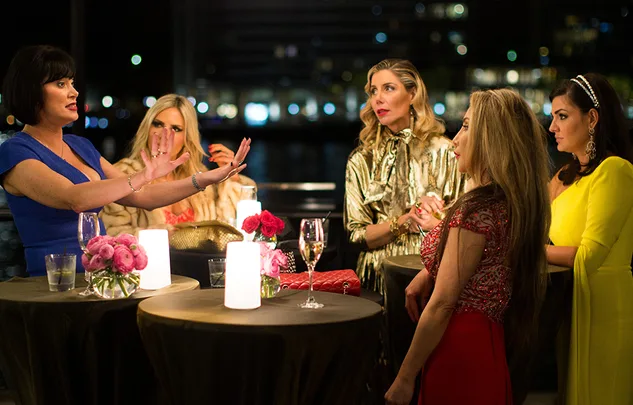Group of women in elegant attire having a conversation at a candlelit table outdoors at night.