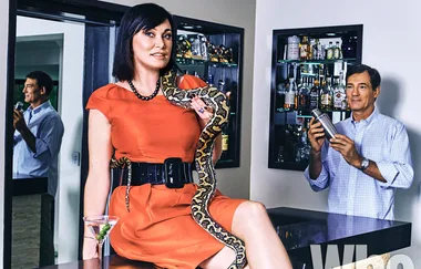 Woman in orange dress with snake draped around her, man shaking cocktail in background, bar with liquor bottles behind.