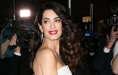 A woman in an elegant dress with wavy hair smiles at a formal event, surrounded by photographers and bright lights.
