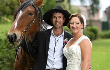 Man in cowboy hat and woman in wedding dress pose with a horse outdoors.