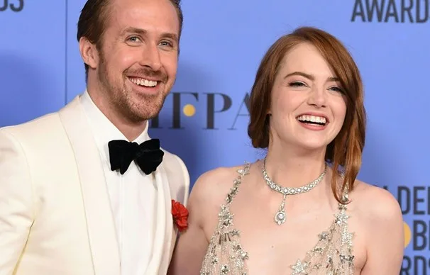 Two smiling people in formal attire at an awards event, against a blue background with logos and text.