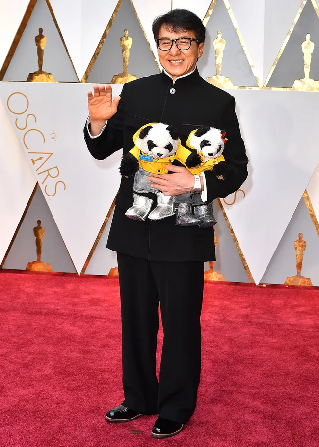Man in black suit holding teddy bears on Oscars red carpet, smiling and waving.
