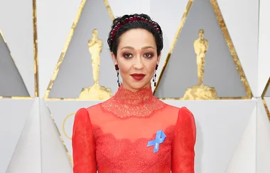 A person in a red lace dress with a blue ribbon on the chest, standing in front of an Oscars-themed backdrop.