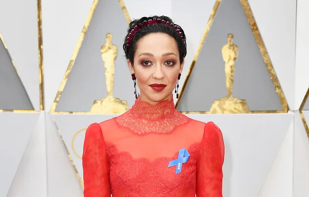 A person in a red lace dress with a blue ribbon on the chest, standing in front of an Oscars-themed backdrop.