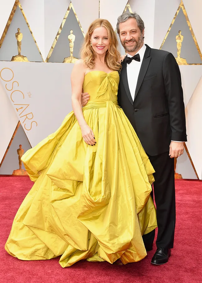 A couple poses on the Oscars red carpet; she wears a yellow gown, he wears a black tuxedo, smiling at the camera.
