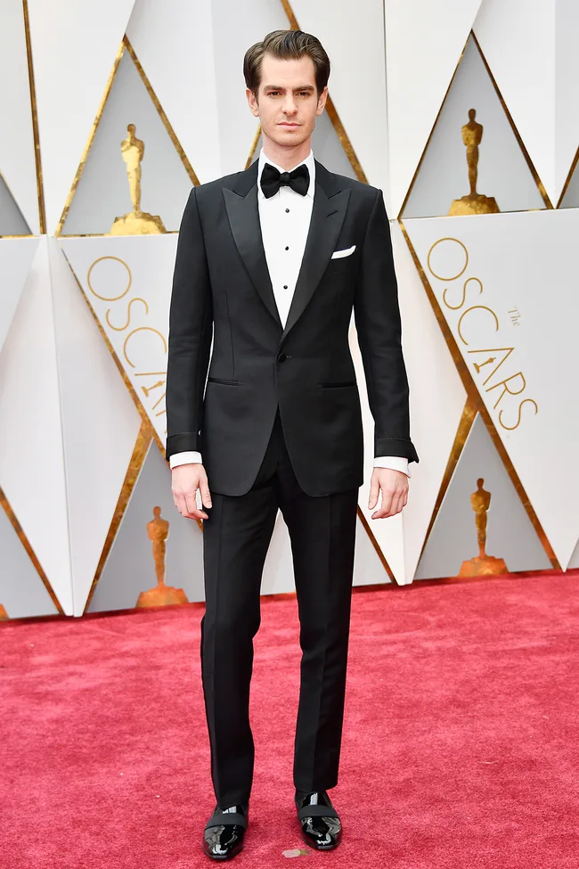 Man in a black tuxedo on the Oscars red carpet, with Oscar statues in the background.