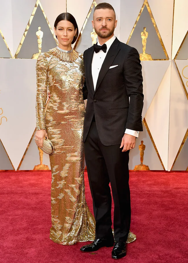A couple in formal attire on a red carpet, with gold geometric backdrop, posing for photos.