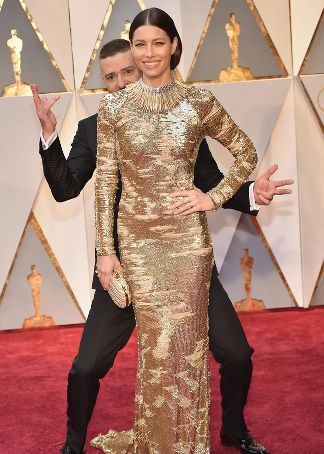 Two people on a red carpet, one in a gold dress posing, the other playfully gesturing behind them.
