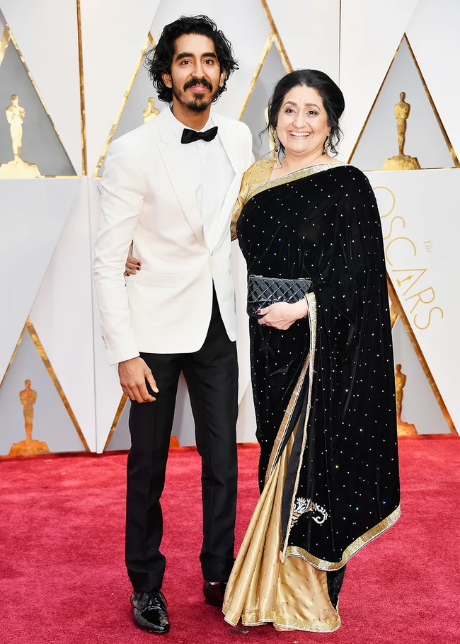 A man in a white tuxedo and a woman in a black saree pose on the red carpet at an award show event.