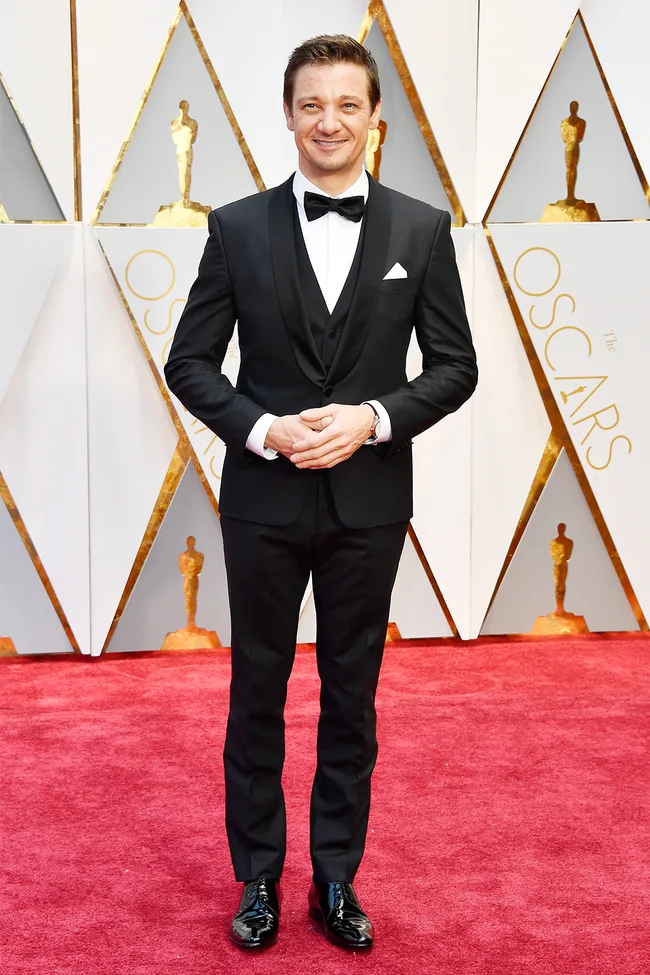 Man in a black tuxedo with bow tie on the Oscars red carpet, smiling and hands clasped.