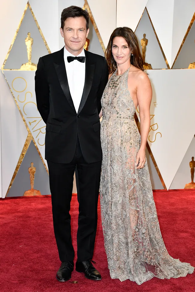 A couple poses on the Oscars red carpet; man in a black tuxedo, woman in a silver lace gown.