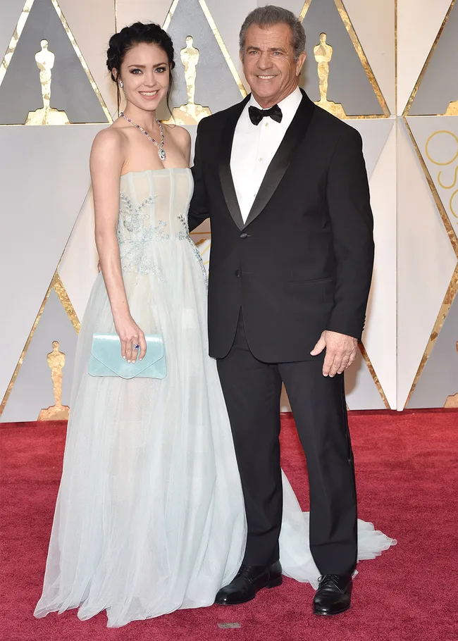 A couple posing on the Oscars red carpet, with the woman in a light blue gown and the man in a black tuxedo.