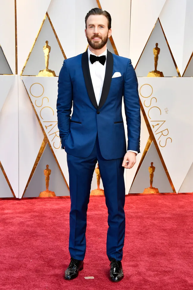 Man in a blue suit with a bow tie on a red carpet at the Oscars.