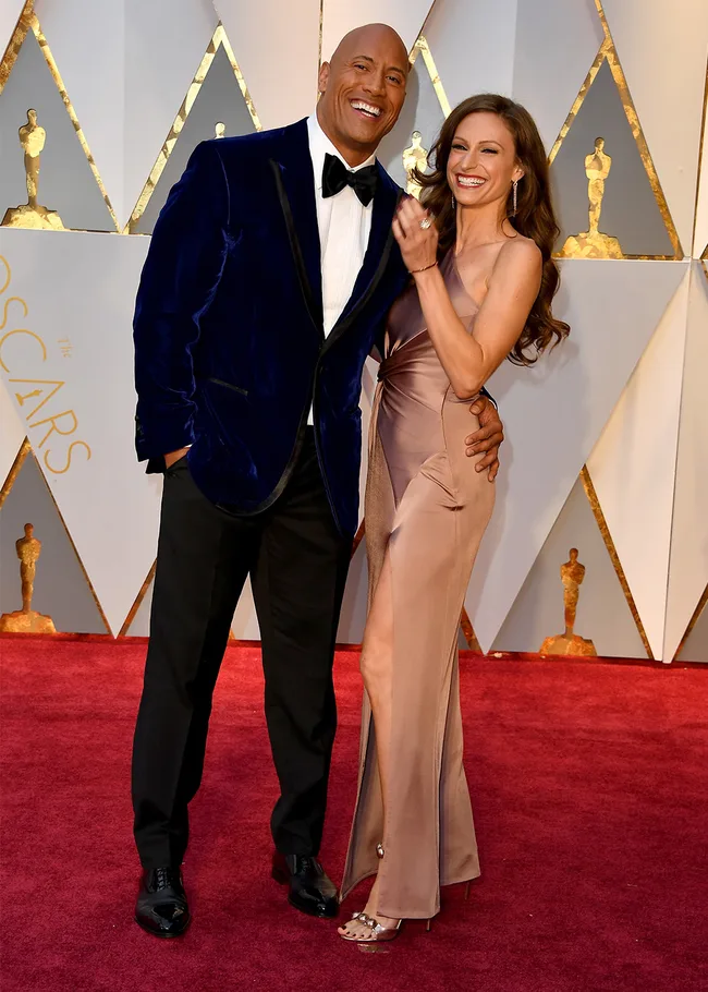 Two people smiling on the Oscars red carpet; man in a suit and woman in a gown.