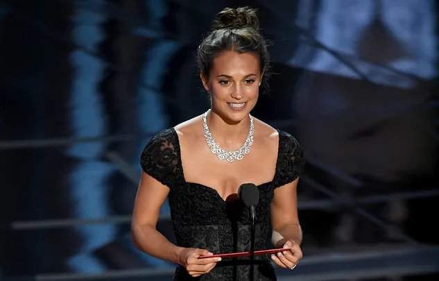 A woman in a black dress and diamond necklace speaks into a microphone at an award ceremony.
