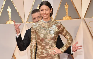Elegant woman in a gold dress on the red carpet, with a man playfully posing behind her.