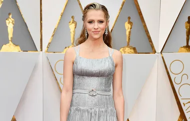 A woman in a silver dress attends a formal event, standing in front of a backdrop with golden Oscar figures.