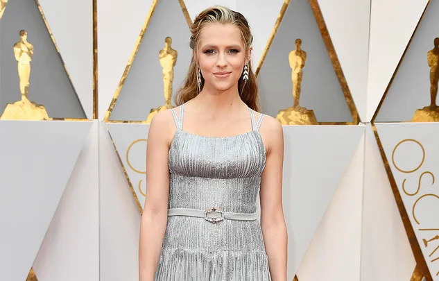 A woman in a silver dress attends a formal event, standing in front of a backdrop with golden Oscar figures.