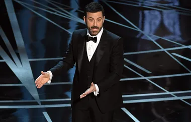 A man in a tuxedo gestures while speaking on a stage with geometric patterns on the floor.