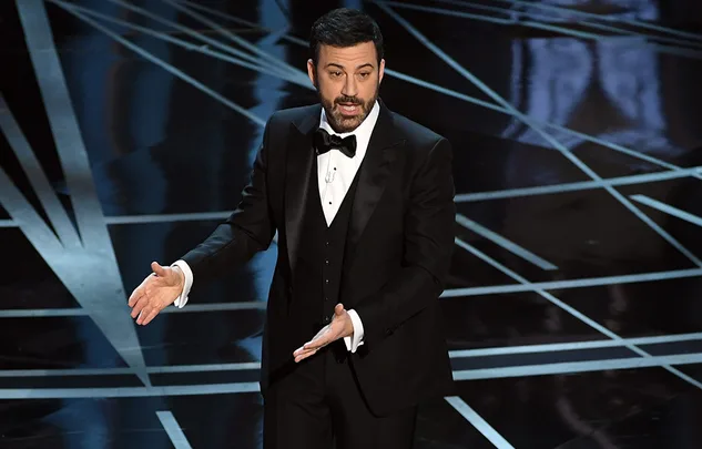 A man in a tuxedo gestures while speaking on a stage with geometric patterns on the floor.