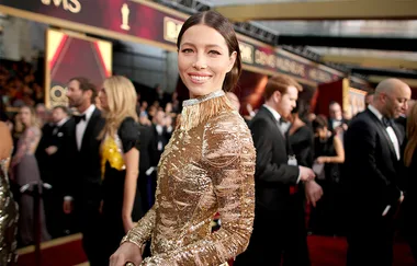 Person in a gold dress smiling on a red carpet at an awards event, with a crowd in the background.