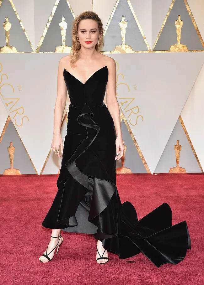 A woman in a black gown poses on the red carpet at the Oscars, with gold Oscar statues in the background.