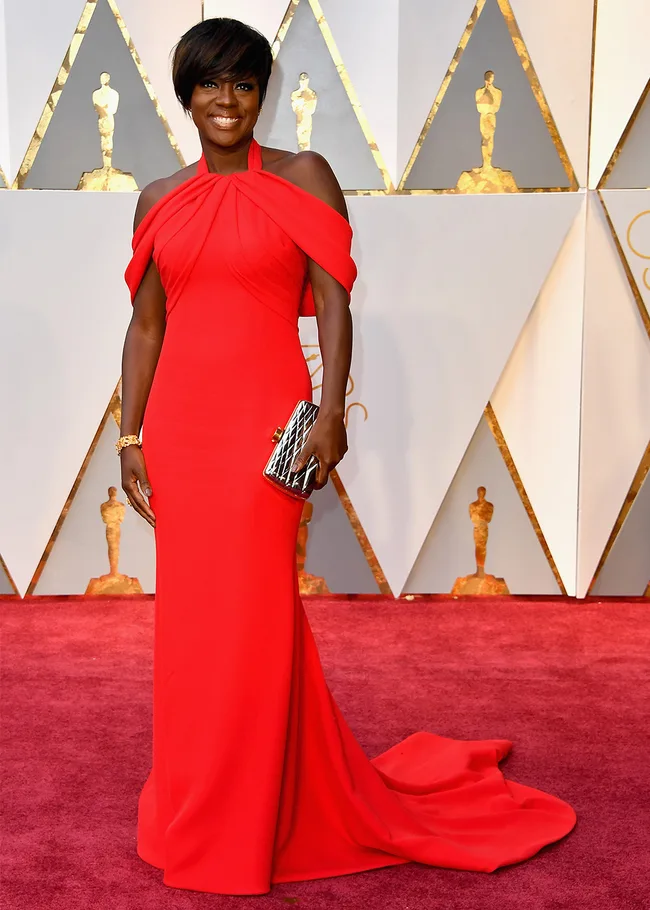 Woman in a red gown holding a clutch on a red carpet with golden Oscar statue backdrop.