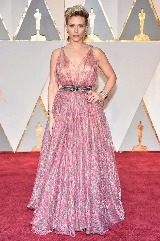 Woman in a pink patterned gown on a red carpet, with backdrop featuring Oscar statuette designs.