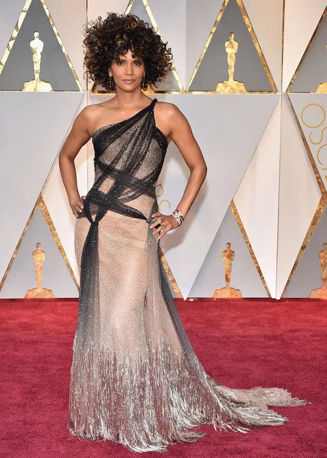Female celebrity in a black and silver gown with curly hair, poses on the Oscars red carpet.
