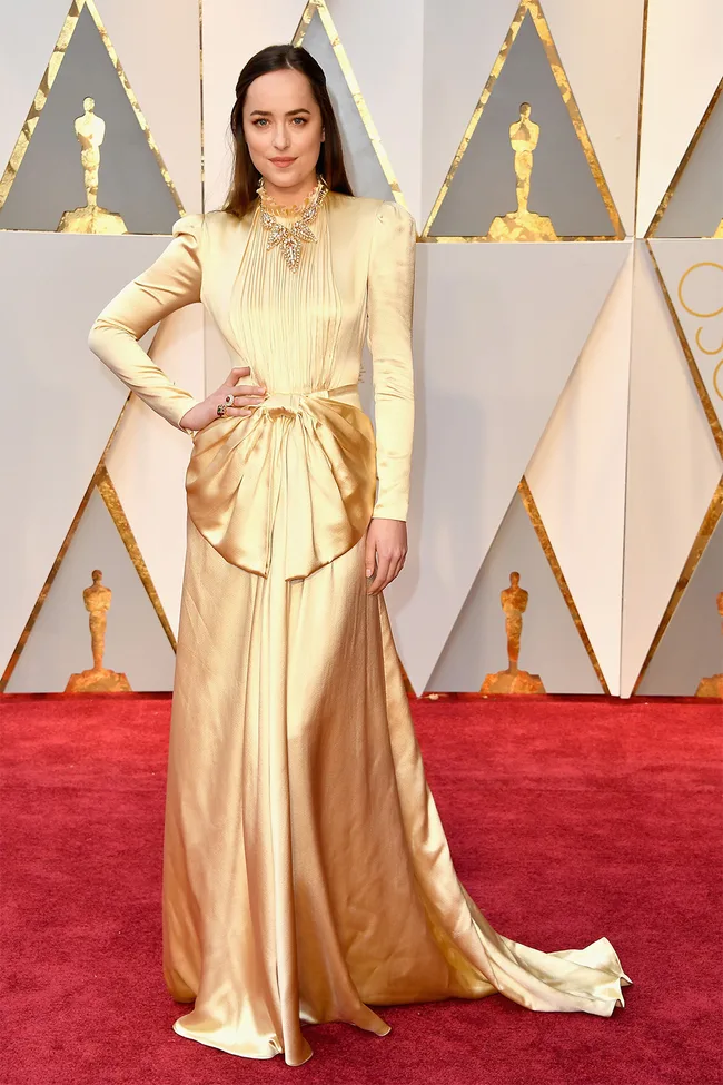 A woman in a long gold gown poses on a red carpet at an event with Oscar-themed decorations in the background.