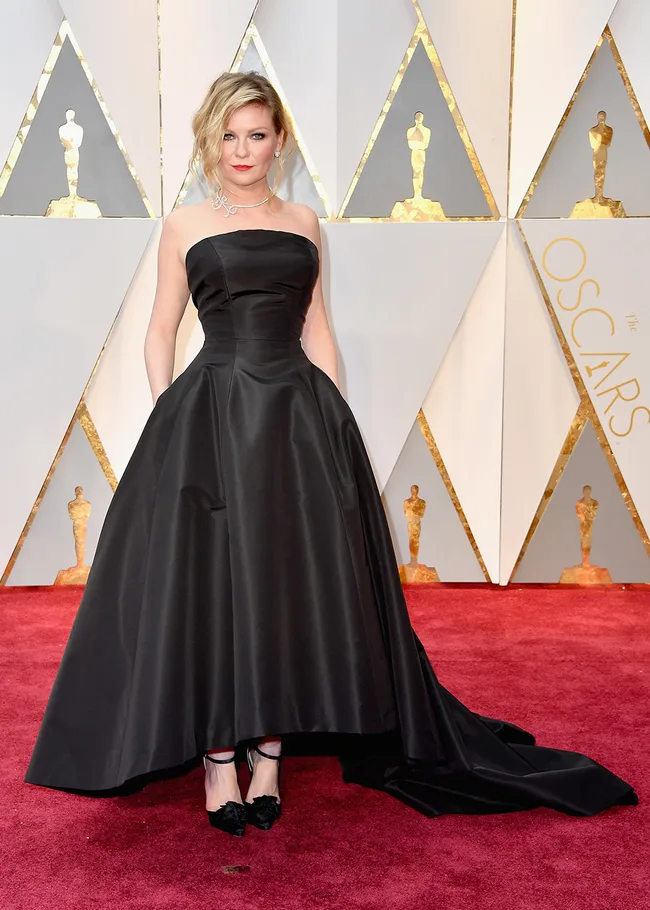 Woman in a strapless black gown poses on the Oscars red carpet with geometric gold and white background.