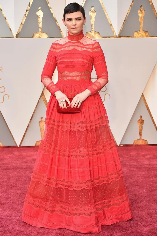 Woman in a red lace gown on a red carpet at an awards event.