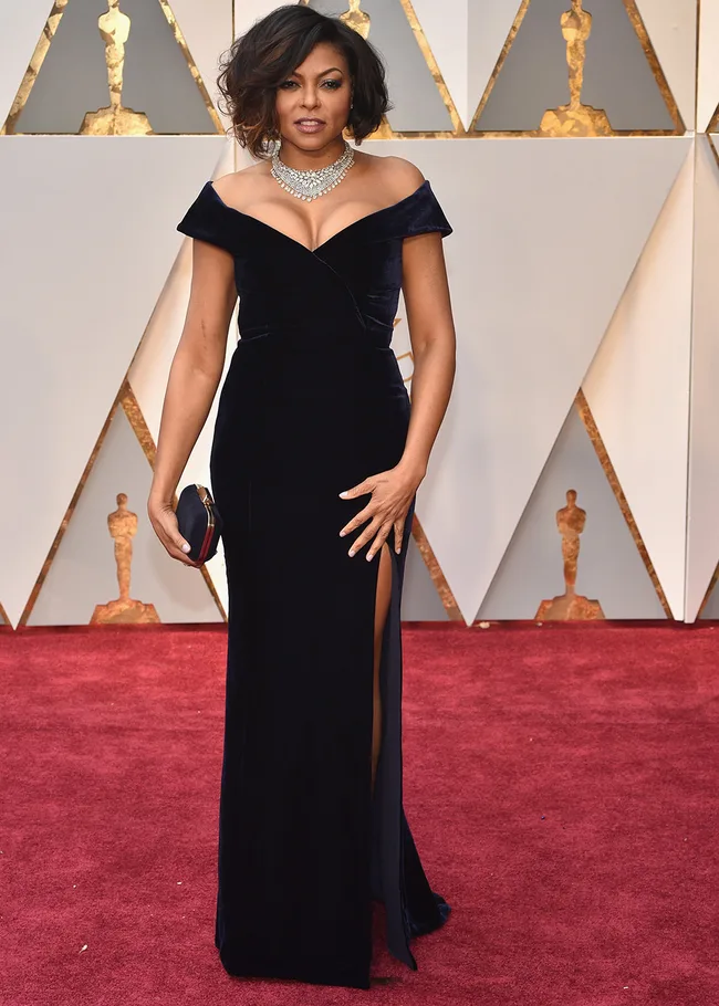 Woman in elegant black gown with side slit, wearing a statement necklace, holding a clutch on a red carpet event.