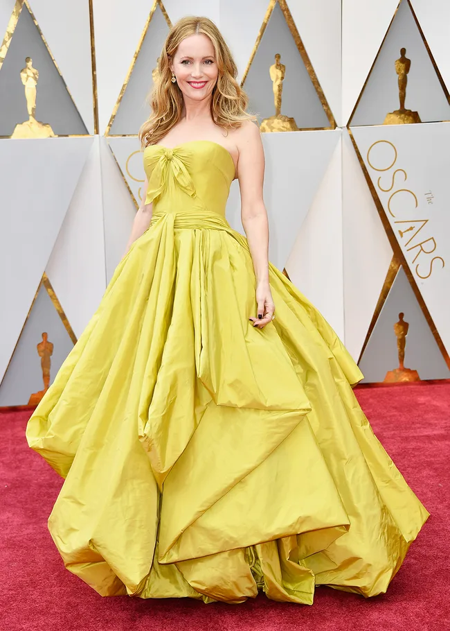 A woman in a bright yellow gown poses on the red carpet at the Oscars, with geometric gold and silver backdrop.