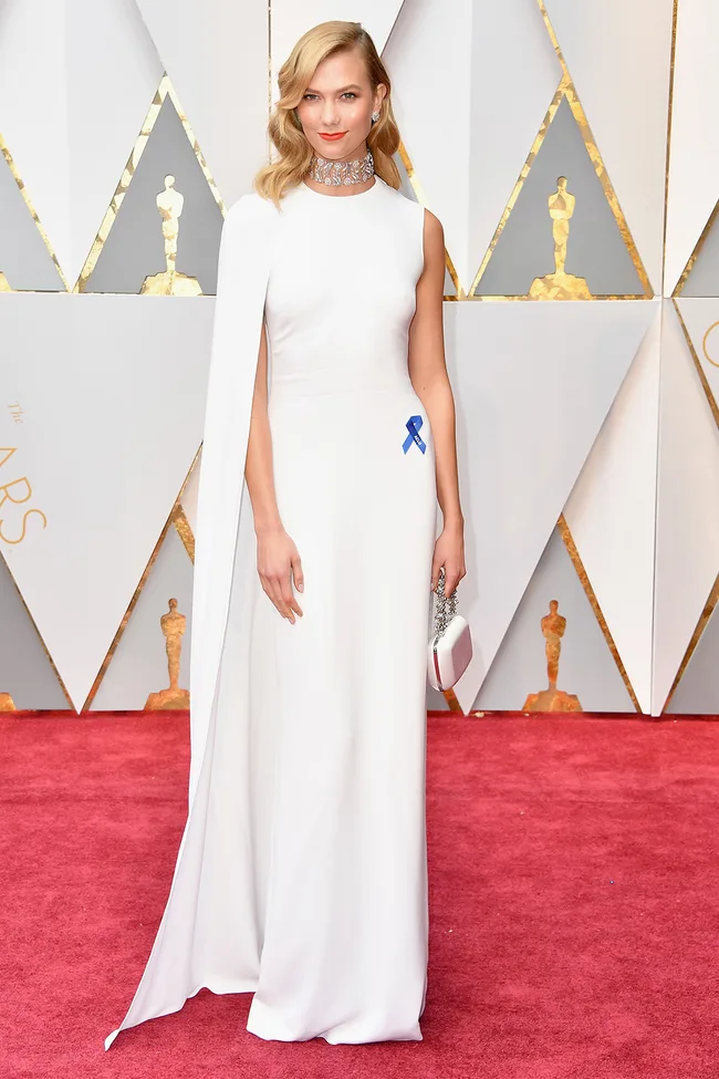 A woman in a white gown with a cape on a red carpet, wearing a blue ribbon and holding a small clutch.