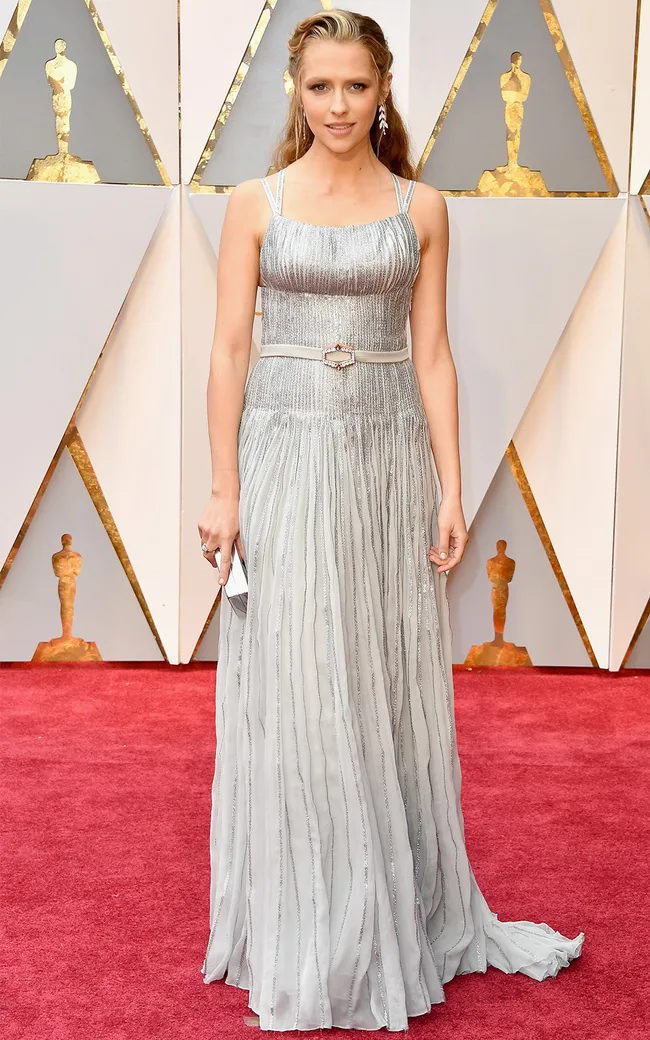 Woman in a silver gown on a red carpet at an awards event, with Oscar statuette backdrop.