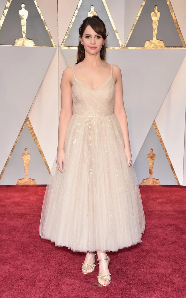 Actress in a light tulle gown on the Oscars red carpet, posing in front of a backdrop with Oscar statuette motifs.