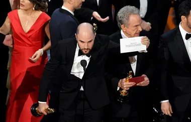 Oscar ceremony mix-up, man holding "Moonlight" Best Picture card, confusion evident on stage, formal attire.