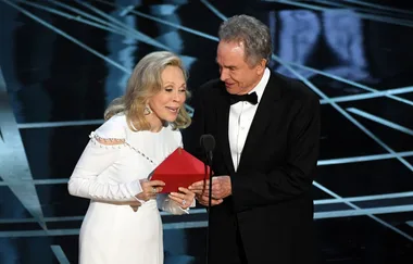 Two people on stage at an awards ceremony, holding a red envelope, smiling and looking at each other.